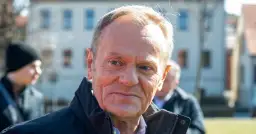 Portret Donalda Tuska w czarnej kurtce, z innymi osobami w tle.