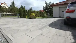 Nowoczesny podjazd z szarej kostki brukowej przed domem z garażem i białym samochodem.