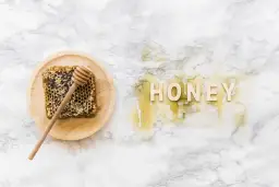 Plaster miodu z drewnianym mieszadełkiem na talerzyku i napis "HONEY" z miodu na marmurowym tle.