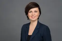 Portret uśmiechniętej kobiety w granatowej marynarce na szarym tle.