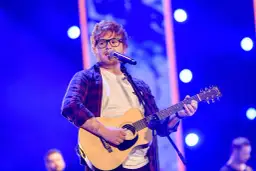 Ed Sheeran w okularach gra na gitarze akustycznej i śpiewa do mikrofonu na niebieskiej scenie.
