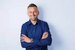 Uśmiechnięty mężczyzna w granatowej koszuli, z rękami skrzyżowanymi na piersi. Idealne zdjecie profilowe meskie.