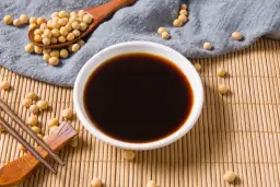 Sos sojowy w miseczce, obok łyżka z ziarnami soi i pałeczki. Dowiedz się, jak zagęścić sos sojowy, by uzyskać idealną konsystencję.