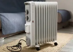 Biały grzejnik olejowy czy elektryczny na kółkach, stojący na dywaniku obok kanapy.