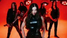 Wokalistka zespołu Arch Enemy w czarnym, błyszczącym stroju, z gitarzystami w tle, na tle czerwonych świateł.