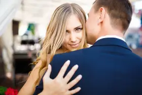 Blondynka z zielonymi oczami patrzy w obiektyw, gdy mężczyzna w granatowym garniturze, numerologiczna 8 mężczyzna, całuje ją w policzek.