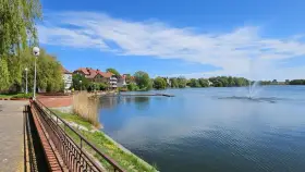 Spokojne jezioro zamkowe z fontanną i drewnianym pomostem, otoczone zielenią i zabudowaniami.