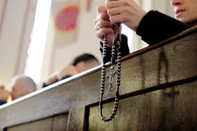 Dłonie trzymające różaniec podczas nabożeństwa. W tle widać innych wiernych, przygotowujących się na odpust.