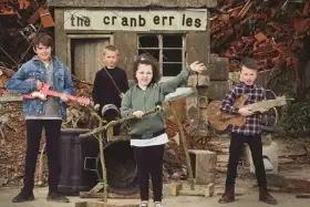 Quatre enfants jouent de la musique devant un panneau "the cranberries". L'un tient une guitare rose, un autre une guitare en bois.