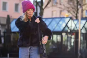 Krótkie futerko stylizacje: blondynka w różowej czapce i czarnym futerku, z jeansami i torebką.