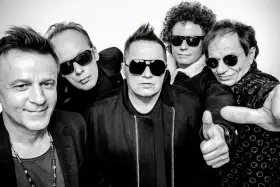 Pięciu mężczyzn w okularach przeciwsłonecznych, gotowych na koncert. Jeden z nich pokazuje kciuk w górę, jakby mówił "t.love hau! hau!".