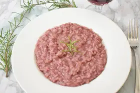 Różowe risotto z prawdziwkami i rozmarynem na białym talerzu, obok widelca i kieliszka wina.