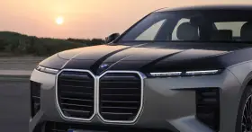Przód luksusowego BMW M7 z podświetlaną atrapą chłodnicy i nowoczesnymi światłami, na tle zachodzącego słońca.