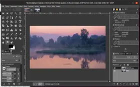 Mglisty poranek nad rzeką, edytowany w **GIMP 2.10**. Widać odbicie drzew w wodzie i wschód słońca.