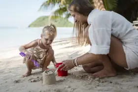 Bambina bionda e mamma giocano con secchiello e paletta sulla sabbia. Un'idea su cosa portare al mare con bambini per divertirsi.