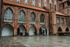 Zamek w Kwidzynie, z ceglanymi arkadami i gotyckimi oknami, skrywa tajemnice. Dwoje ludzi z parasolem spaceruje po mokrym dziedzińcu.