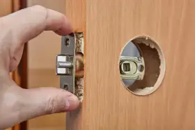 Dłoń montuje mechanizm zamka w drewnianych drzwiach. Dowiedz się, ile kosztuje wymiana zamka w drzwiach.