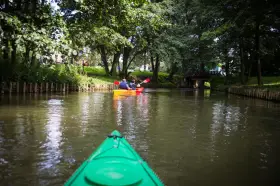 Dwie osoby płyną kajakiem po spokojnej rzece Radunia, otoczonej zielenią i drzewami.