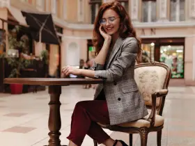 Elegancka kobieta w okularach i marynarce w kratę, prezentująca stylizacje z bordowymi spodniami, siedzi przy stoliku w kawiarni.