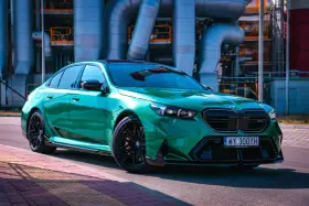 Błyszczące, zielone BMW M5 G90 z czarnymi felgami stoi na tle przemysłowych rur.