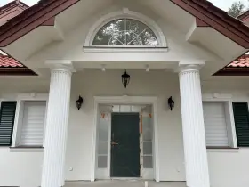 Elegancki pilaster zdobi fasadę domu, wspierając dach nad wejściem. Obok okna i drzwi, tworzy klasyczną kompozycję.