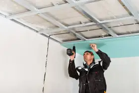 Un obrero instala un **pladur techo** con una taladradora. La estructura metálica ya está fijada al techo original.