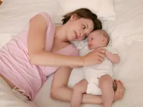 Mama patrzy z miłością na swoje 5 tygodniowe dziecko leżące obok niej na łóżku.
