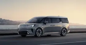 Szary Volvo EM90 jedzie wzdłuż wybrzeża o zachodzie słońca.