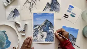 Une main peint une peinture montagne facile, avec des tubes de peinture et des nuanciers bleus à proximité.