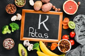 Tablica z napisem "K Potas" otoczona owocami i warzywami bogatymi w potas, jak banany, awokado, pomidory, ziemniaki, brokuły, rodzynki, morele, kiwi, migdały, pestki dyni. Pomaga zapobiegać hipokaliemii.