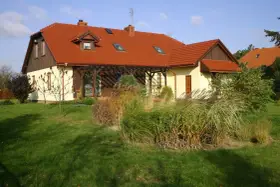 Dom z czerwonym dachem i drewnianym naczółkiem, otoczony zielonym trawnikiem i ozdobnymi trawami.