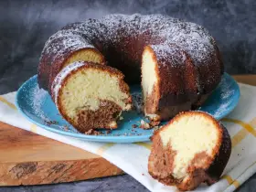 Saftiger weltbester Marmorkuchen, mit Puderzucker bestäubt, auf blauem Teller. Ein Stück ist abgetrennt und zeigt die köstliche Marmorierung.