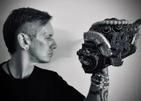 Mężczyzna w czarnym t-shircie patrzy na tradycyjną maskę. Czerń koloru podkreśla tajemniczość sceny.