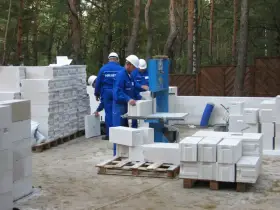 Budowa domu z bloczków betonowych. Pracownicy w niebieskich kombinezonach i białych kaskach przygotowują materiał. Widać stosy bloczków, piłę do cięcia i palety.