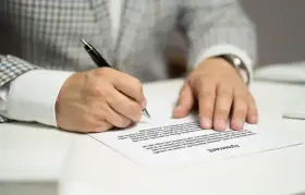 Mężczyzna w eleganckim garniturze podpisuje umowę sprzedaży mieszkania. Dokument leży na białym stole.