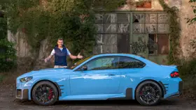 Mężczyzna prezentuje błękitne BMW M2 G87 przed zniszczonym budynkiem.