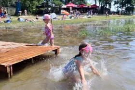 Dwie dziewczynki pluskają się w wodzie przy drewnianym pomoście. W tle plażowicze i drzewa. Wakacje nad jeziora lubelskie.