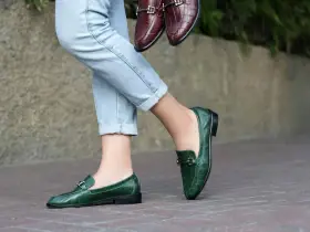 Un mocassino verde con abito elegante, perfetto per un look sofisticato. Un altro paio marrone è tenuto in mano.