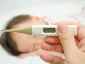 Cyfrowy termometr pokazuje 39.5°C. W tle widać śpiące dziecko. Zastanawiasz się, ile czasu mierzyć temperaturę pod pachą?