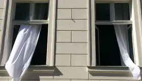 Weiße Gardinen wehen aus zwei offenen Fenstern eines hellen Gebäudes. Die Frage "Gardinen waschen wieviel Grad?" beschäftigt die Hausfrau.