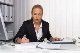 Kobieta w biurze pracuje nad dokumentami, używając kalkulatora i długopisu. Na biurku leżą stosy papierów i segregatorów, a w tle widać regał z dokumentacją. Może przygotowuje weksle.