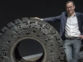 Wim Delvoye pose à côté d'une roue ornée de dragons, une œuvre d'art monumentale.