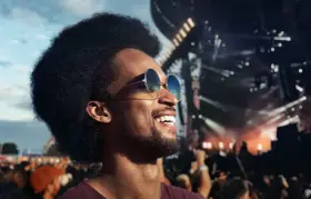 Homme souriant à un concert, portant des lunettes de soleil et un bouchon d'oreille pour protéger son audition.