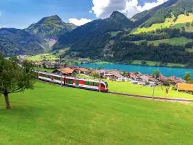A red and white train travels along the **Golden Pass Line Lucerne to Interlaken**, passing a village by a turquoise lake, with lush green mountains in the background.