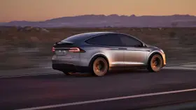 Srebrny Tesla X pędzi przez pustynny krajobraz o zachodzie słońca. Zastanawiasz się nad jego ceną?