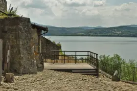 Kamienna wieża i drewniany taras z widokiem na spokojne jezioro dobczyckie. W oddali zalesione wzgórza.