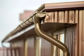 Brązowa rynna z widocznym spadkiem, odprowadzająca wodę z dachu. Widać fragmenty brązowej elewacji i okna.