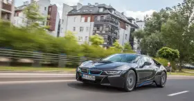 Czarny BMW i8 pędzi ulicą, mijając nowoczesne budynki i zielone drzewa.