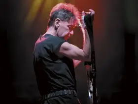 Muzyk z zespołu KSU z gitarą na scenie, w czarnej koszulce i z charakterystyczną fryzurą.