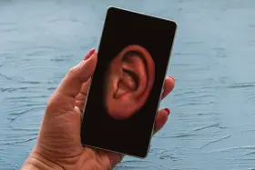 Dłoń trzyma smartfon z uchem na ekranie. Czy Twój telefon jest na podsłuchu? Sprawdź to!